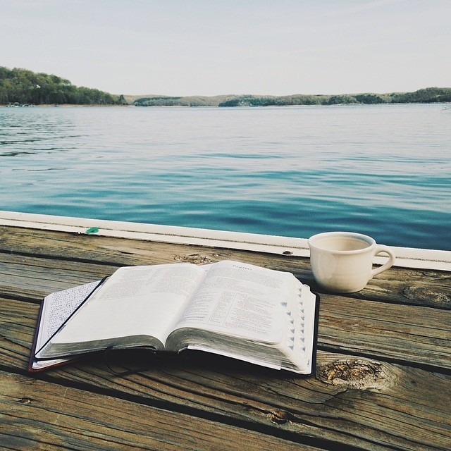 Open book on a weathered wooden dock beside a calm blue lake, with a white coffee mug nearby.