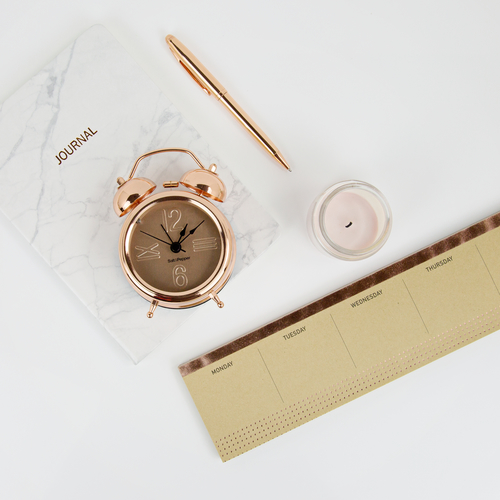 Flat lay of a marble journal labeled 'JOURNAL', a rose-gold alarm clock, a gold pen, a small candle, and a beige weekly planner on a white surface.