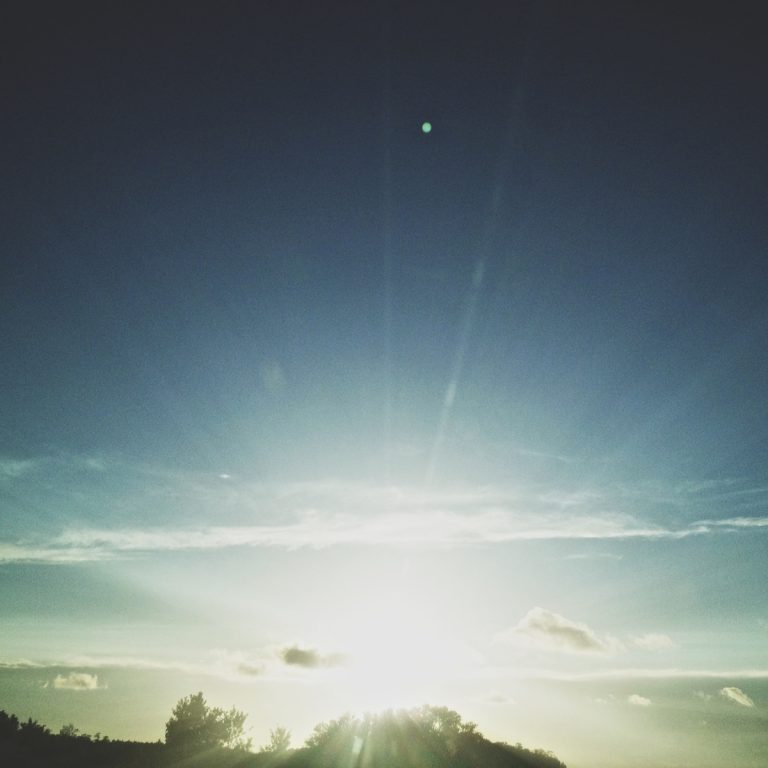 Sunrise over a treeline with bright sun and lens flare streaks across the soft sky.