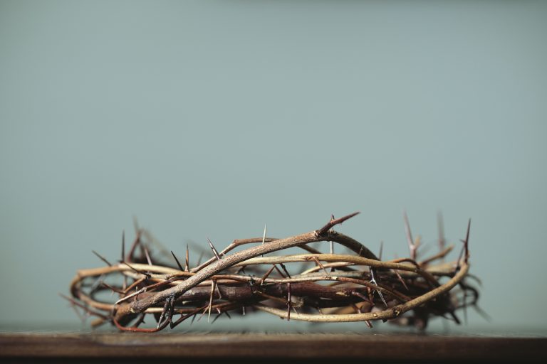 Close-up of a crown of thorns made from dried branches resting on a wooden surface with a pale blue background, symbolic religious imagery.