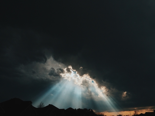 Beams of sunlight breaking through dark storm clouds onto a suburban horizon at sunset.