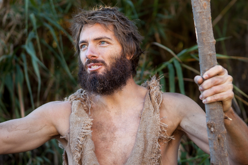 Bearded man in tattered burlap clothing grips a wooden staff in a dense jungle backdrop
