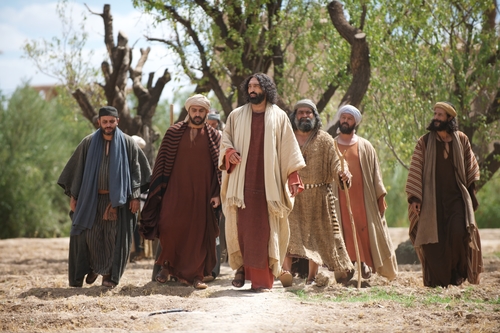 Group of men in biblical-era robes walking together on a dirt path under trees in sunlight.