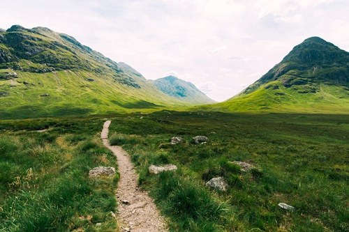 Dirt path winding through green moorland toward distant mountains under a pale sky.